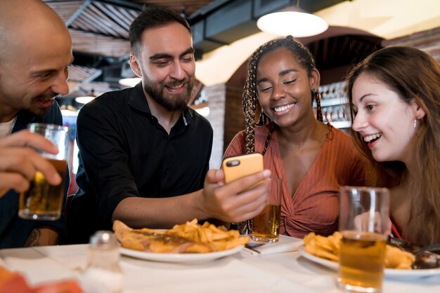 Grupo de amigos diversos que utilizan un teléfono móvil mientras disfrutan de una comida juntos en un restaurante. Concepto de amigos.