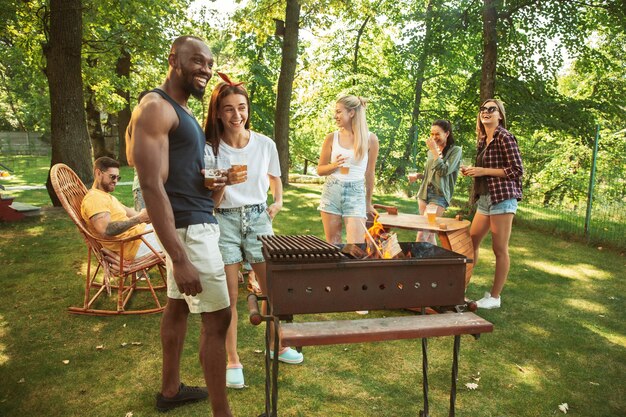 Grupo de amigos felices con cerveza y fiesta de barbacoa en un día soleado. Descansar juntos al aire libre en un claro del bosque o en el patio trasero. Celebrar y relajarse, reír. Estilo de vida de verano, concepto de amistad.