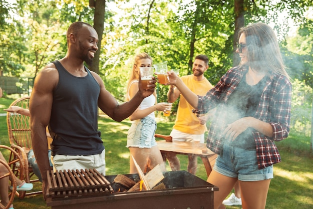 Grupo de amigos felices con cerveza y fiesta de barbacoa en un día soleado. Descansar juntos al aire libre en un claro del bosque o en el patio trasero. Celebrar y relajarse, reír. Estilo de vida de verano, concepto de amistad.