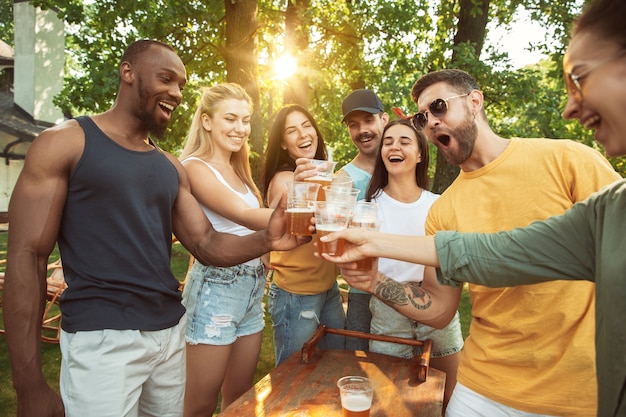 Grupo de amigos felices con cerveza y fiesta de barbacoa en un día soleado. Descansar juntos al aire libre en un claro del bosque o en el patio trasero