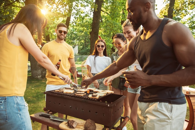 Foto gratuita grupo de amigos felices con cerveza y fiesta de barbacoa en un día soleado. descansar juntos al aire libre en un claro del bosque o en el patio trasero