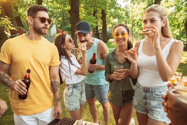 Grupo de amigos felices con cerveza y fiesta de barbacoa en un día soleado. Descansar juntos al aire libre en un claro del bosque o en el patio trasero