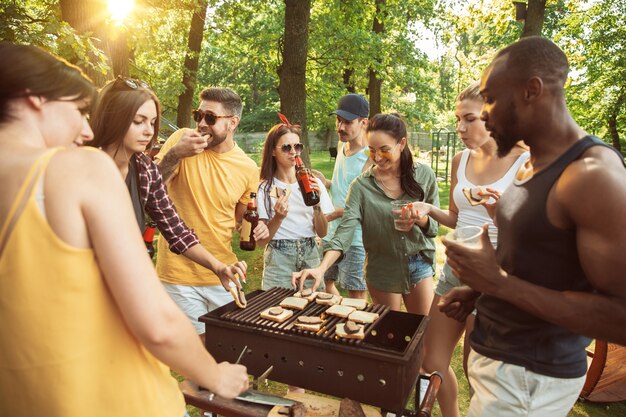 Grupo de amigos felices con cerveza y fiesta de barbacoa en un día soleado. Descansar juntos al aire libre en un claro del bosque o en el patio trasero