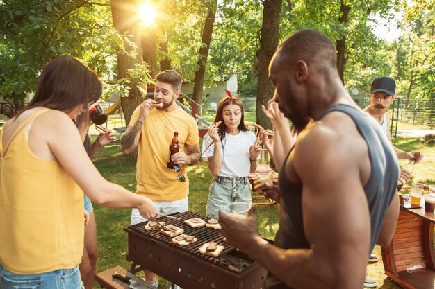 Grupo de amigos felices con cerveza y fiesta de barbacoa en un día soleado. Descansar juntos al aire libre en un claro del bosque o en el patio trasero