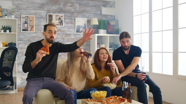 Grupo de amigos felices después de que su equipo de fútbol favorito gane el campeonato. Amigos sentados en el sofá comiendo pizza y papas fritas.