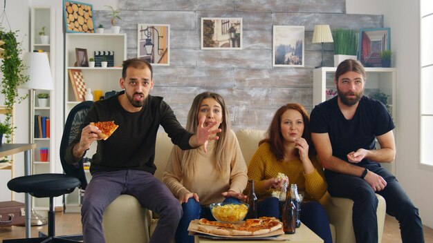 Grupo de amigos felices después de que su equipo de fútbol favorito gane el campeonato. Amigos sentados en el sofá comiendo pizza y papas fritas.