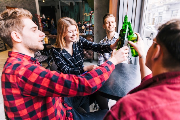 Grupo de amigos felices sentados alrededor de la mesa de madera tostando las botellas de cerveza verde en pub