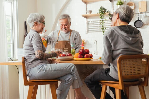 Grupo de amigos mayores que tienen una fiesta en el interior cocinando y conversando positivamente en un senior daycareasian Senior masculino de pie en la cocina y relajándose en casa mientras come una receta de comida saludable