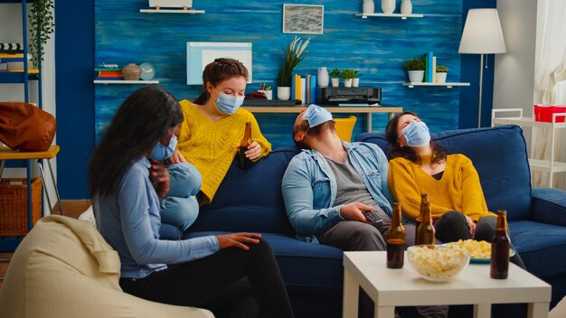 Grupo de amigos multiétnicos viendo un programa de comedia en la televisión riendo con una mascarilla para prevenir la infección con covid 19, durante la pandemia global divirtiéndose sentado en el sofá manteniendo la distancia social