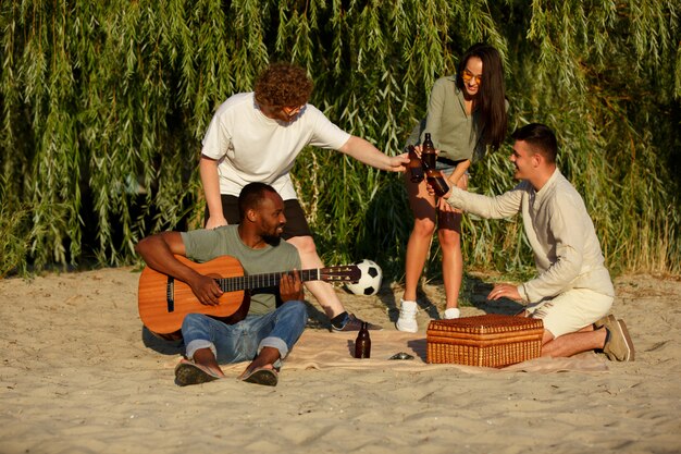 Grupo de amigos tintineando botellas de cerveza durante un picnic en la playa. Estilo de vida, amistad, diversión, fin de semana y concepto de descanso.