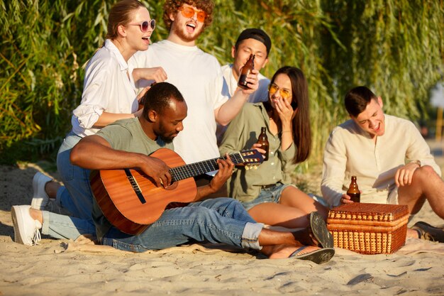 Grupo de amigos tintineando botellas de cerveza durante un picnic en la playa. Estilo de vida, amistad, diversión, fin de semana y concepto de descanso.