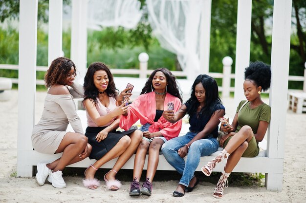 Grupo de cinco chicas afroamericanas relajándose en una hermosa cabaña junto a la piscina al lado de un resort de lujo y mirando teléfonos móviles