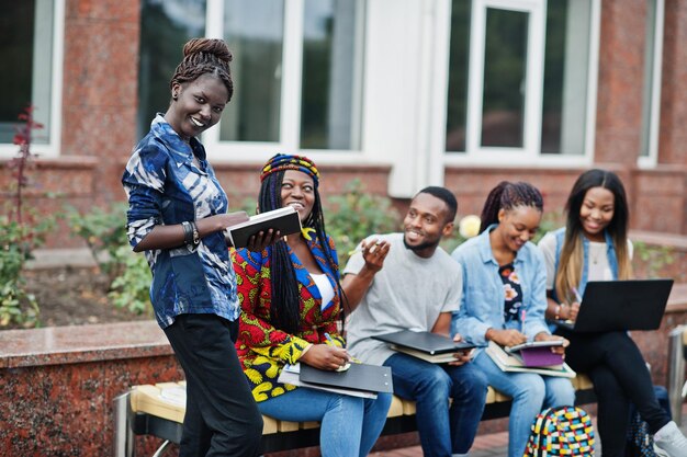 Grupo de cinco estudiantes universitarios africanos que pasan tiempo juntos en el campus en el patio de la universidad Amigos afro negros que estudian en un banco con artículos escolares computadoras portátiles cuadernos