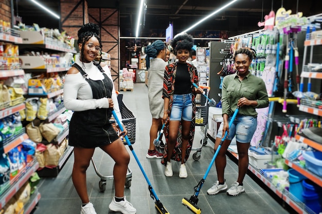 Grupo de cinco mujeres africanas con trapeador de polvo y cubo divirtiéndose en el departamento de artículos de limpieza del hogar en el supermercado