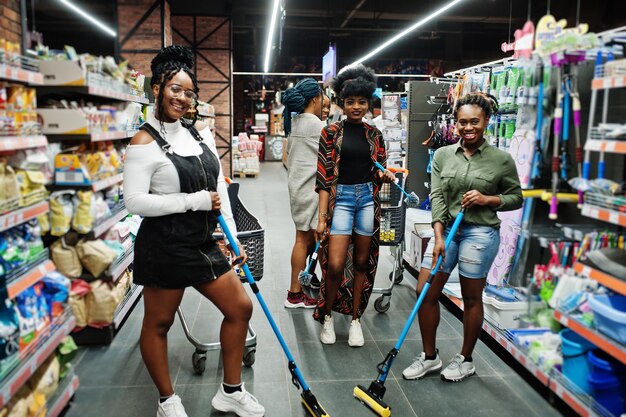 Grupo de cinco mujeres africanas con trapeador de polvo y cubo divirtiéndose en el departamento de artículos de limpieza del hogar en el supermercado