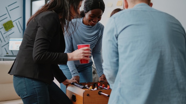 Grupo de colegas jugando al futbolín después del trabajo, bebiendo cerveza y comiendo pizza en la oficina. Gente disfrutando de actividades divertidas después de horas con fútbol y bebidas alcohólicas.