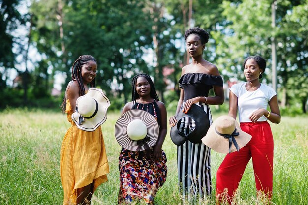 Grupo de cuatro hermosas mujeres afroamericanas usan sombrero de verano pasando tiempo en la hierba verde en el parque