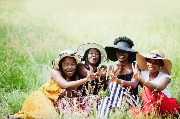 Grupo de cuatro hermosas mujeres afroamericanas usan sombrero de verano sentado en la hierba verde en el parque