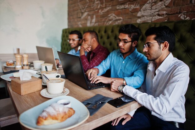 Grupo de cuatro hombres del sur de Asia posaron en una reunión de negocios en un café Los indios trabajan juntos con computadoras portátiles usando varios dispositivos conversando