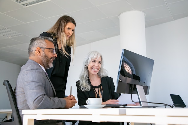 Grupo empresarial feliz viendo la presentación y riendo. Profesionales sentados juntos en el lugar de trabajo, mirando el monitor de la computadora y riendo. Concepto de comunicación empresarial o trabajo en equipo