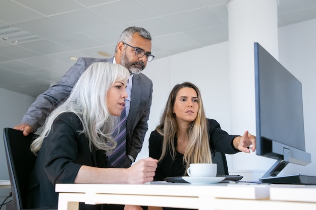 Grupo empresarial viendo la presentación en el monitor de la PC y discutiendo el proyecto, sentado en el lugar de trabajo con una taza de café y apuntando a la pantalla. Concepto de comunicación empresarial