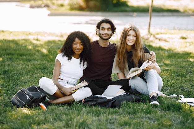 Grupo de estudiantes internacionales sonrientes sentados juntos en un césped en el parque en la Universidad. Niñas africanas y caucásicas y niño indio hablando al aire libre
