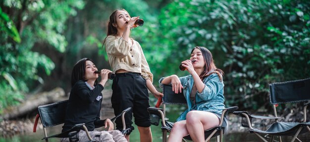 Grupo hermosas mujeres asiáticas amigas viajeras relajándose en sillas de campamento en el arroyo Están animando y bebiendo cerveza durante el campamento hablando con diversión y felices juntas