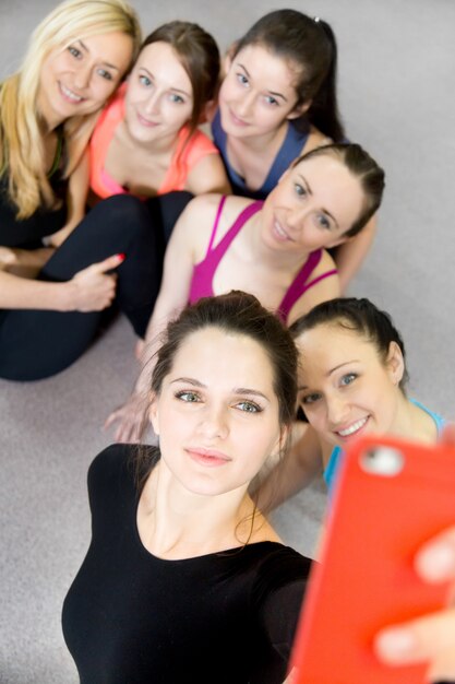 Grupo de hermosas niñas deportivas posando para selfie, auto-retrato con smartphone rojo en gimnasio deportivo