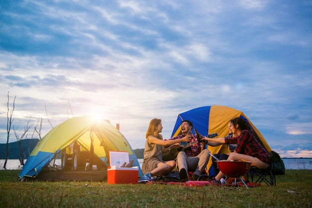 Foto gratuita grupo de hombre y mujer disfrutar de picnic y barbacoa de camping en el lago con tiendas de campaña en el fondo. joven mujer de raza mixta asiática y el hombre. las manos de los jóvenes tostando y animando botellas de cerveza.