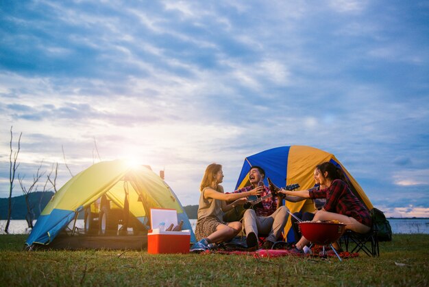 Grupo de hombre y mujer disfrutar de picnic y barbacoa de camping en el lago con tiendas de campaña en el fondo. Joven mujer de raza mixta asiática y el hombre. Las manos de los jóvenes tostando y animando botellas de cerveza.