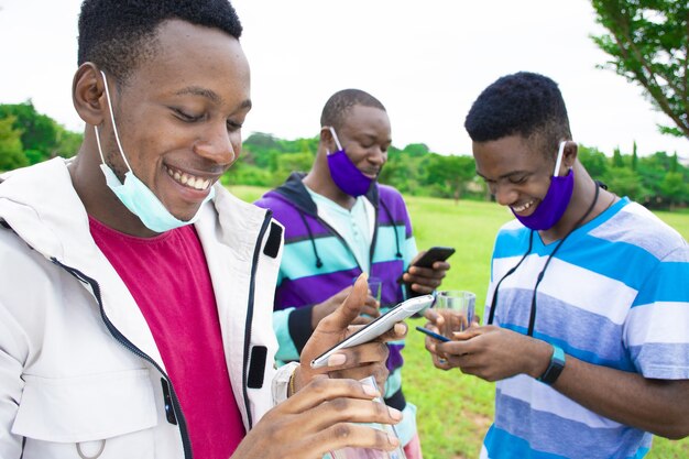 Grupo de jóvenes amigos africanos con mascarillas usando teléfonos mientras el distanciamiento social en un parque