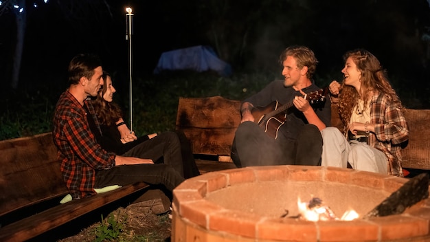 Un grupo de jóvenes amigos felices cerca de una fogata en glamping, noche. Dos hombres y mujeres. Tocar la guitarra