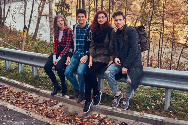 Grupo de jóvenes amigos con mochilas sentados en la barandilla cerca de la carretera con un hermoso bosque y un río al fondo. Viajes, caminatas, concepto.