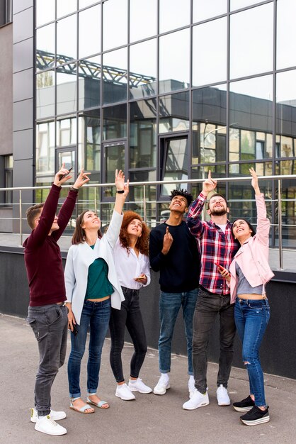 Grupo de jóvenes amigos de pie en la calle apuntando hacia arriba frente a un edificio moderno