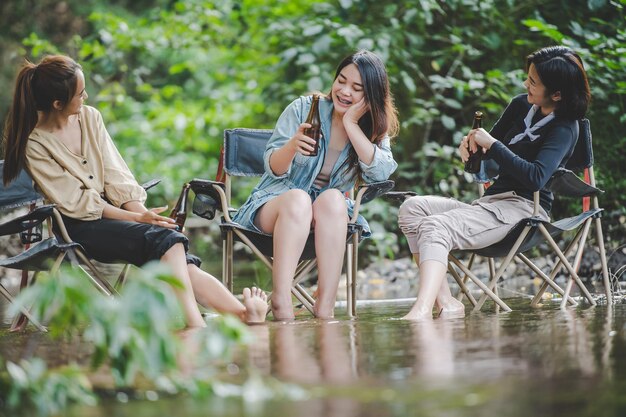 Un grupo de jóvenes asiáticas beben cerveza en sus sillas y se mojan los pies en el arroyo mientras acampan en el parque natural Disfrutan hablando y riéndose juntas