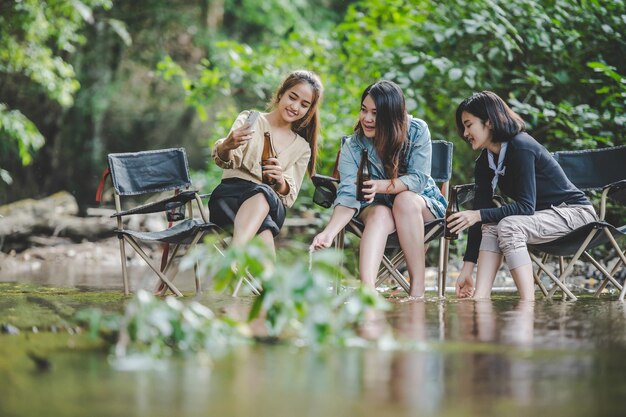 Un grupo de jóvenes asiáticas beben cerveza en sus sillas y se mojan los pies en el arroyo mientras acampan en el parque natural Disfrutan hablando y riéndose juntas