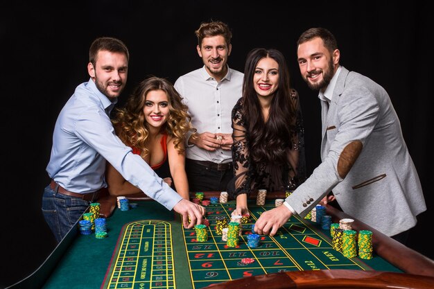 Grupo de jóvenes detrás de la mesa de la ruleta sobre fondo negro. Los jóvenes están apostando en el juego. Jugadores mirando a la cámara.