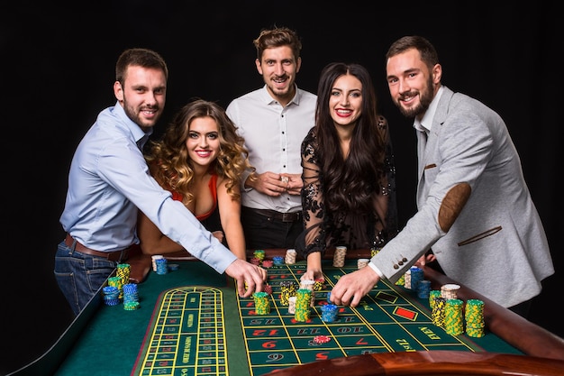 Grupo de jóvenes detrás de la mesa de la ruleta sobre fondo negro. Los jóvenes están apostando en el juego. Jugadores mirando a la cámara.