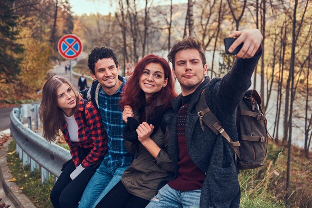 Grupo de jóvenes excursionistas con mochilas sentados en barandilla cerca de la carretera y haciendo selfie con un hermoso bosque y río en el fondo.