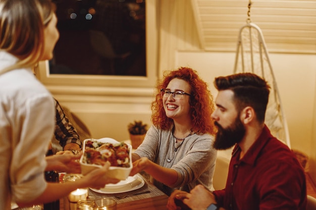 Grupo de jóvenes felices cenando juntos en la mesa de comedor El foco está en la mujer pelirroja tomando comida de su amiga