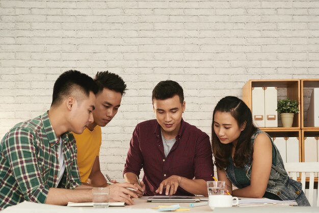 Grupo de jóvenes hombres y mujeres asiáticos de pie juntos alrededor de la mesa y mirando la tableta