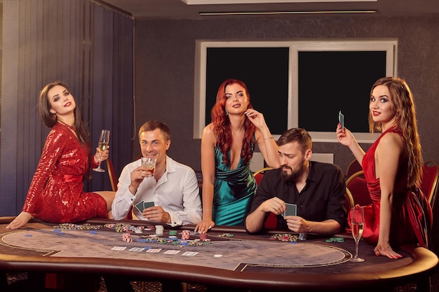 Foto gratuita un grupo de jóvenes jugadores posan en una mesa de póquer en el casino y miran la cámara. los jóvenes están haciendo apuestas esperando una gran victoria. juegos de azar por dinero. juegos de fortuna.