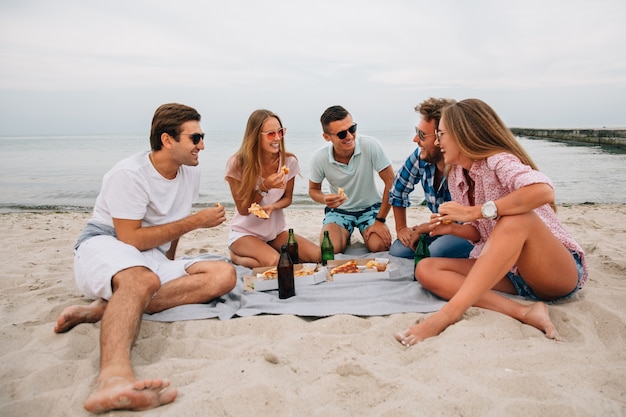 Grupo de jóvenes sonrientes chicos y chicas descansando juntos en la playa, sentado cerca del mar