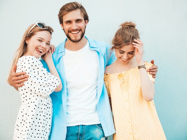 Foto gratuita grupo de jóvenes tres elegantes amigos posando en la calle. hombre de moda y dos chicas lindas vestidas con ropa casual de verano. modelos sonrientes divirtiéndose cerca de la pared. mujeres alegres y chico al aire libre