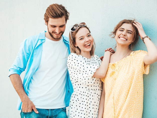 Foto gratuita grupo de jóvenes tres elegantes amigos posando en la calle. hombre de moda y dos chicas lindas vestidas con ropa casual de verano. modelos sonrientes divirtiéndose cerca de la pared. mujeres alegres y chico al aire libre