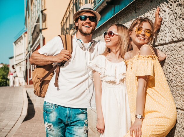 Grupo de jóvenes tres elegantes amigos posando en la calle. Hombre de moda y dos chicas lindas vestidas con ropa casual de verano. Modelos sonrientes divirtiéndose en gafas de sol. Mujeres alegres y chico al aire libre