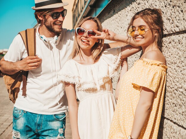 Grupo de jóvenes tres elegantes amigos posando en la calle. Hombre de moda y dos chicas lindas vestidas con ropa casual de verano. Modelos sonrientes divirtiéndose en gafas de sol. Mujeres alegres y chico al aire libre