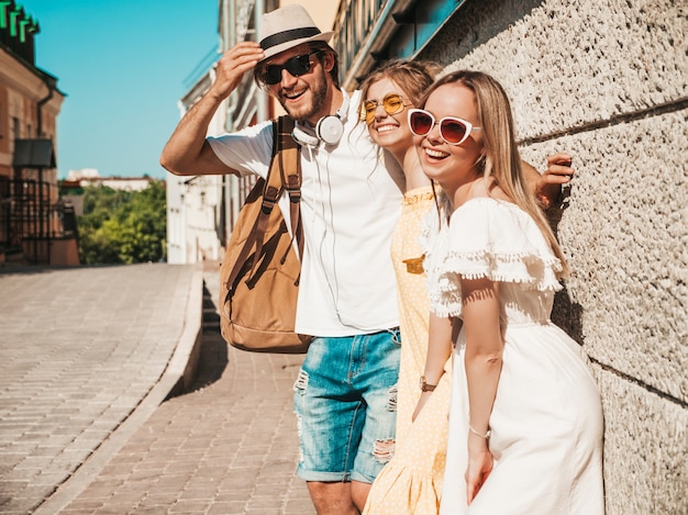 Grupo de jóvenes tres elegantes amigos posando en la calle. Hombre de moda y dos chicas lindas vestidas con ropa casual de verano. Modelos sonrientes divirtiéndose en gafas de sol. Mujeres alegres y chico al aire libre