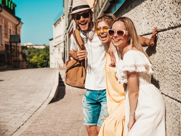 Grupo de jóvenes tres elegantes amigos posando en la calle. Hombre de moda y dos chicas lindas vestidas con ropa casual de verano. Modelos sonrientes divirtiéndose en gafas de sol. Mujeres alegres y chico al aire libre