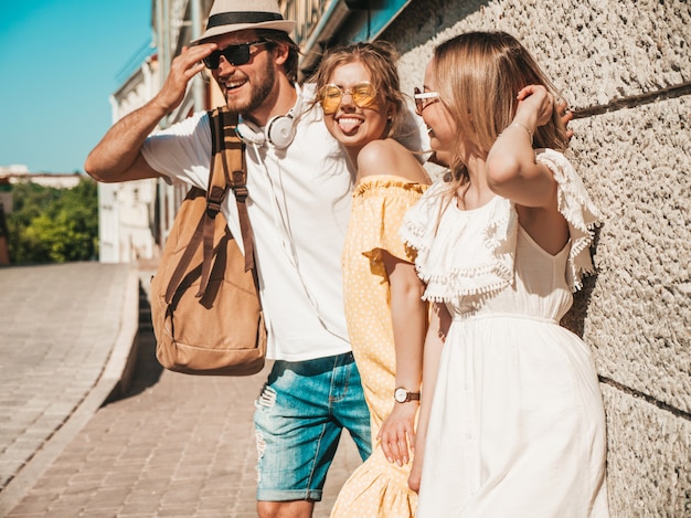 Foto gratuita grupo de jóvenes tres elegantes amigos posando en la calle. hombre de moda y dos chicas lindas vestidas con ropa casual de verano. modelos sonrientes divirtiéndose en gafas de sol. mujeres alegres y chico al aire libre